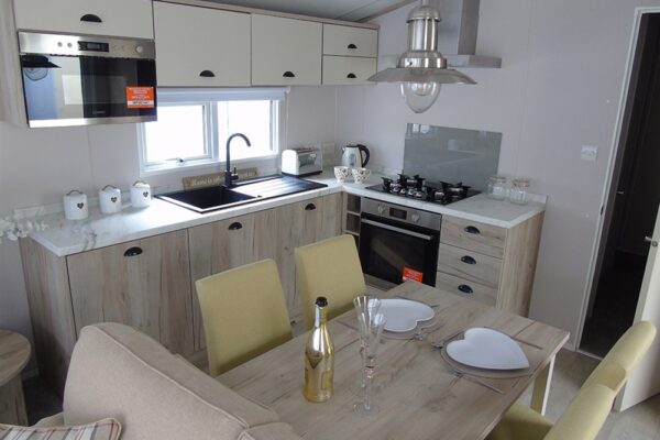 Modern kitchen with light wood cabinets, stainless oven, and a dining table set for a meal with heart-shaped plates and champagne flutes on a pale wood table.
