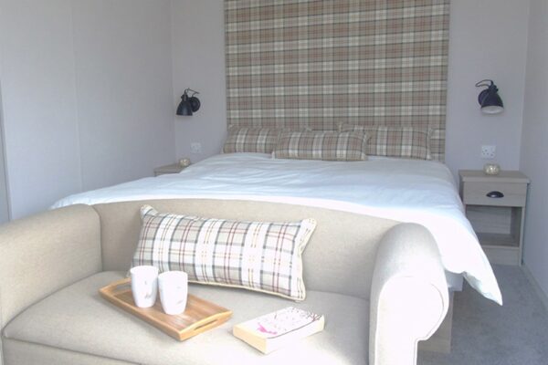 A beige sofa with plaid cushion in front of a white bed with a plaid headboard and matching pillows, two nightstands with lamps on either side, and a wooden tray with two mugs and a book on the sofa.