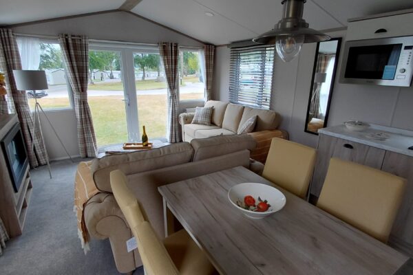 Bright caravan living area with beige sofa, plaid curtains, and a marble dining table; wine bottle and glasses on a tray by the window.