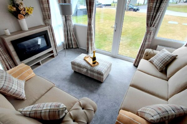 Cozy living room with beige plaid sofas and cushions, an ottoman tray holding a champagne bottle and glasses, near large glass doors.