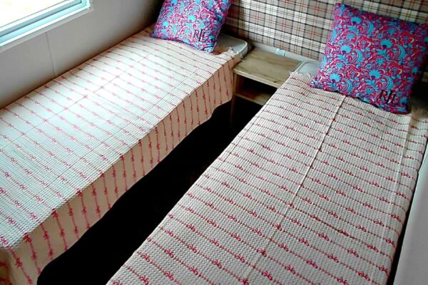 Two single beds with plaid headboards and cream-red striped bedspreads, colorful floral pillows, and a small wooden nightstand between them in a cozy room.