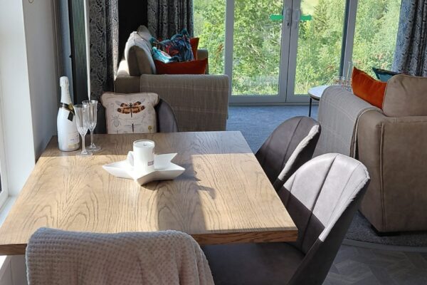 Bright dining area with a wooden table, beige chairs, a champagne bottle and glasses, and a countryside view through glass doors.