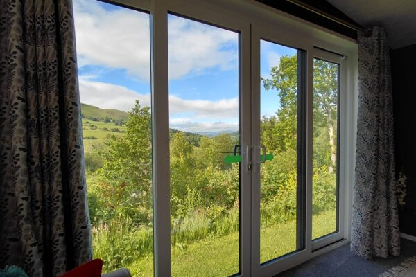 Interior view of a large glass window with white frames and floral-patterned curtains, showing a sunny countryside scene outside with hills and trees.