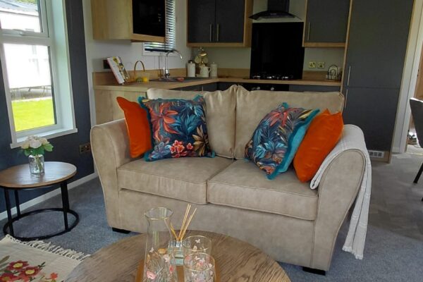 Open-plan living space with a beige sofa and colorful tropical cushions, facing a wood-and-dark cabinet kitchen backdrop.