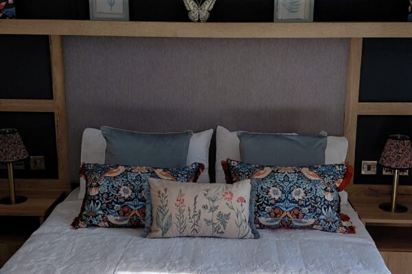 A neatly made bed with a padded headboard, blue and floral throw pillows, and a round wooden tray holding teacups on a white quilted duvet in a stylish bedroom.