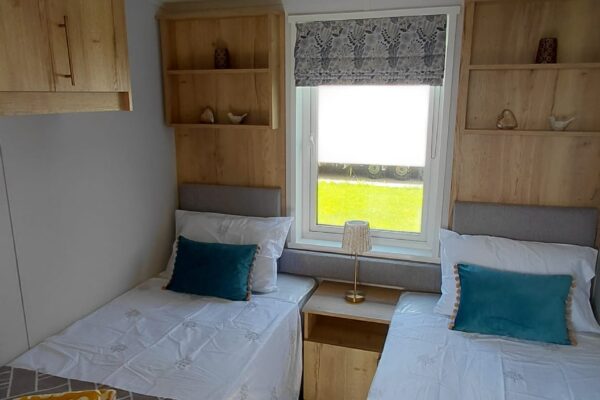 Twin bedroom with two single beds, teal pillows, and wooden wall-mounted shelves surrounding a central window with a grey blind.