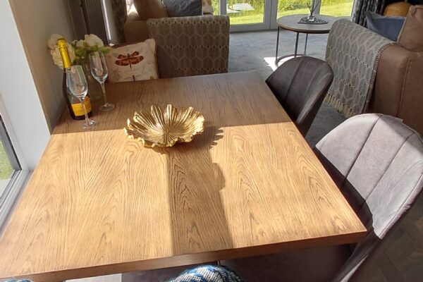 Wood dining table with a decorative shell centerpiece, a wine bottle and two glasses, in a sunlit living area with glass doors opening to a garden.