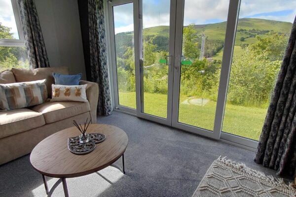 Living room with a beige sofa and patterned cushions facing large glass doors opening to green hills outside; blue floral curtains frame the view.