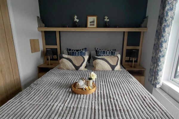 A neatly made bed with striped bedding, two patterned pillows, and a wooden headboard shelf above the bed in a hotel-like room.