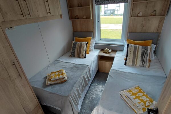 Twin bedroom with two single beds, gray bedding, and mustard pillows, separated by a small wooden nightstand under a window; wood-panel walls and built-in shelves.