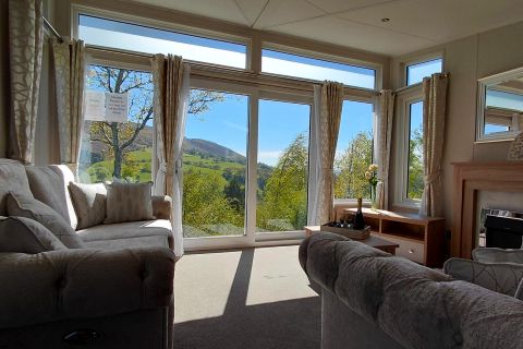 Bright living room with two beige sofas and large floor-to-ceiling windows revealing a green hillside and blue sky outside.