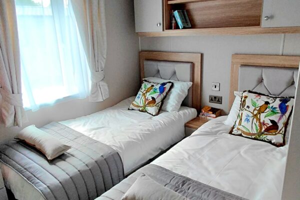 Twin bedroom with two single beds, white bedding, gray quilted runners, and decorative bird pillows under light wood headboards.