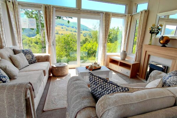 Bright living room with beige sofas, patterned cushions, and a panoramic window view of rolling countryside outside the glass doors and windows.