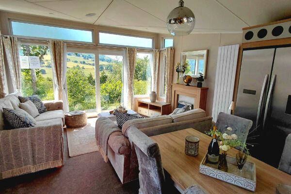 Cozy living room with cream sofas and patterned cushions, facing large windows that overlook rolling countryside. A wooden coffee table sits between them near a fireplace and decorative items.