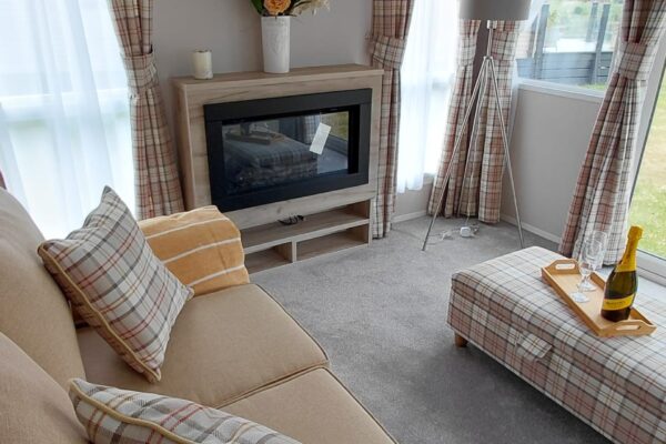 Cozy living room with a beige plaid sofa, plaid curtains, and a fireplace topped with a vase of yellow and orange flowers.