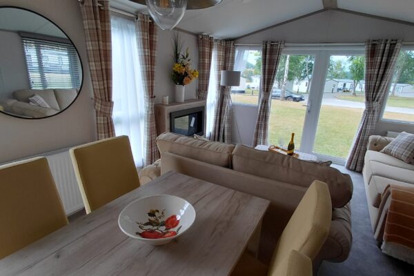 Cozy living area in a mobile home: beige sofas, plaid curtains, round wall mirror, and a fireplace; dining table with a decorative bowl and a wine bottle nearby.